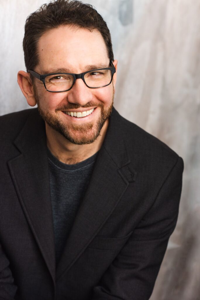 Jeremy Sortore, a white man wearing dark clothing and glasses, smiling in front of a light grey background.